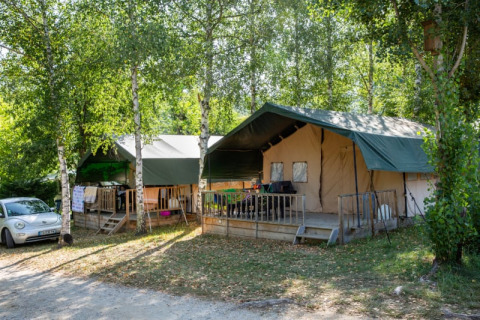 Two safari tents with green roofs surrounded by leafy trees, with a car parked nearby on a campsite.