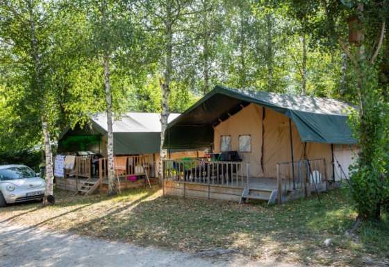 Two safari tents with green roofs surrounded by leafy trees, with a car parked nearby on a campsite.