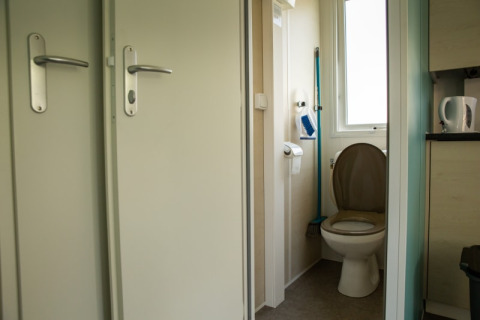 Baño pequeño con inodoro, papel higiénico, escobilla y ventana en Sun Lodge, Camping Les Bouleaux, Francia.
