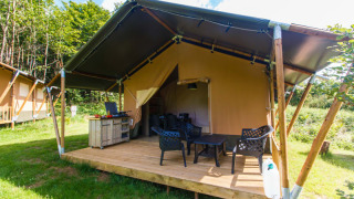 Safari tent with private terrace, outdoor chairs, and table, set on a grassy campsite surrounded by greenery.