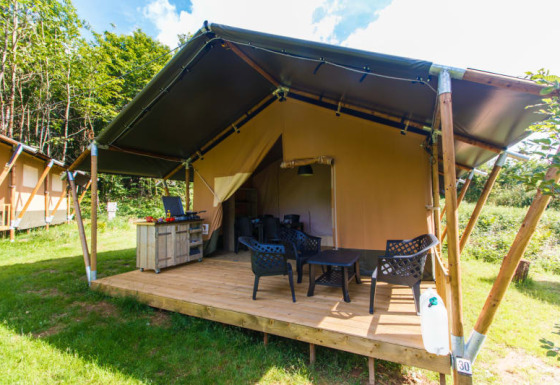 Tienda safari con terraza privada, sillas y mesa al aire libre, ubicada en un camping rodeado de naturaleza.