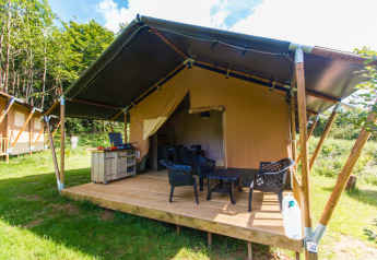 Safari tent with private terrace, outdoor chairs, and table, set on a grassy campsite surrounded by greenery.