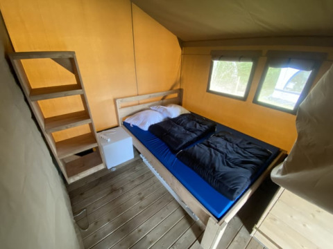 Vista interior de una tienda safari con cama doble, mini nevera y suelo de madera en Camping Jagtveld.