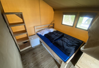 Vista interna di tenda safari con letto matrimoniale, mini frigo e pavimento in legno al Camping Jagtveld.