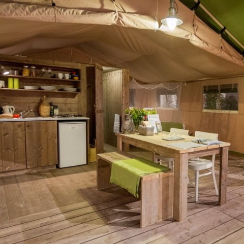 Interno di tenda safari con arredi in legno, cucina e tavolo da pranzo a Tenuta Tredici Ulivi, Italia.