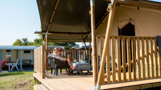 Tenda safari con servizi igienici, due persone rilassate sulla veranda in legno coperta immersa nella natura.