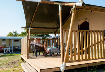 Tenda safari con servizi igienici, due persone rilassate sulla veranda in legno coperta immersa nella natura.