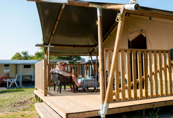 Tienda safari con baños privados, dos personas relajándose en una terraza de madera cubierta al aire libre.