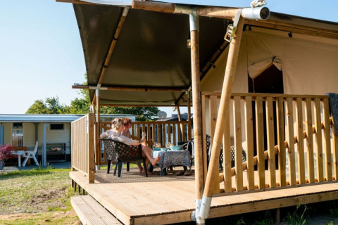 Safari-Zelt mit Sanitäranlagen, zwei Menschen entspannen auf der überdachten Holzterrasse im Freien.