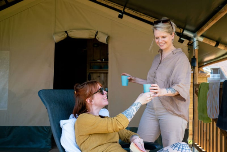 Twee vrouwen genieten van drankjes voor een safaritent met eigen sanitair bij Camping Tussen de Diepen, Nederland.