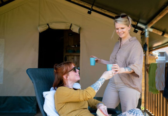 Twee vrouwen genieten van drankjes voor een safaritent met eigen sanitair bij Camping Tussen de Diepen, Nederland.