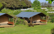 To hyggelige glamping telte med terrasse står på en grøn græsplæne, omgivet af buske og træer.