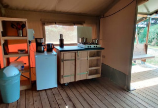 Área de cocina interior en una tienda glamping con suelo de madera, estufa, nevera y vista exterior.