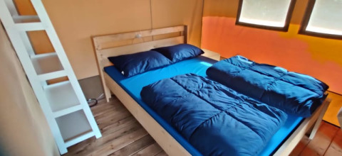 Interior de una tienda safari con cama de madera, ropa de cama azul, ventanas y una escalera blanca.