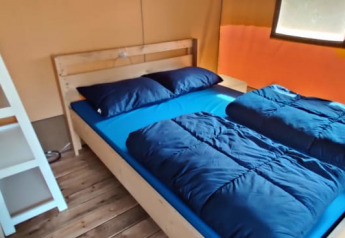 Interno di tenda safari con letto in legno, biancheria da letto blu, finestre e scala bianca.