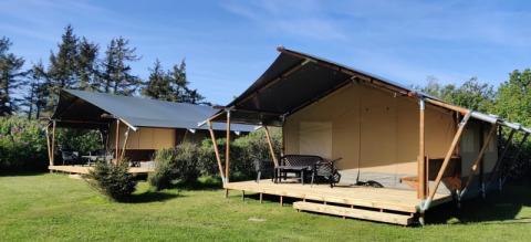 Deux tentes safari avec terrasse en bois placées sur une pelouse, entourées d’arbres sous un ciel bleu.