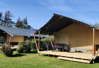 Two safari tents with wooden decks are set on grassy lawns, surrounded by trees under a blue sky.