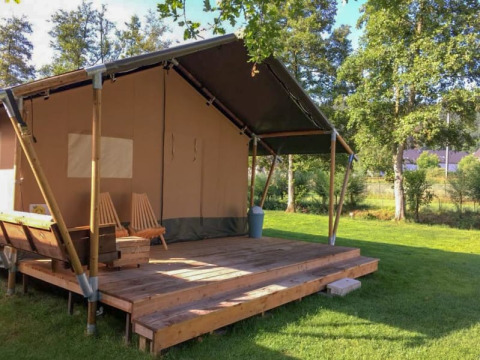 Tienda de safari con terraza de madera y sillas, situada en un césped verde rodeado de árboles.