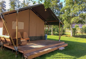 Safaritent met houten veranda en stoelen, geplaatst op een groen gazon omringd door bomen.