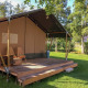 Safari tent with wooden deck and chairs, set on a green lawn surrounded by trees, in daylight.