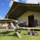Safari telt lodge med træ picnicbord og en grå hund, der ligger på græsset under en blå himmel.