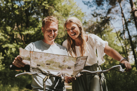 Twee mensen glimlachen terwijl ze samen een kaart lezen op hun fietsen in het bos bij Beerze Bulten.