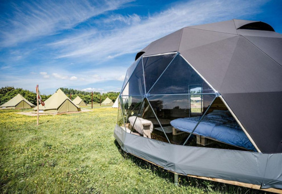 Maison nature Cast Away Dome avec dôme géodésique et tentes, sur prairie fleurie sous ciel bleu clair.