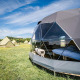 Glamping på Cast Away Dome naturhus med kuppeltelt og telte på en eng under en klar blå himmel.