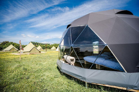 Maison nature Cast Away Dome avec dôme géodésique et tentes, sur prairie fleurie sous ciel bleu clair.