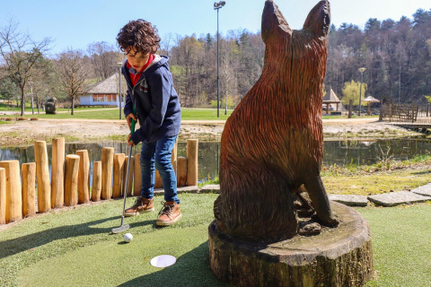 Niño jugando minigolf junto a una gran estatua de zorro de madera en Cosy Cabins en Limburg, Bélgica.