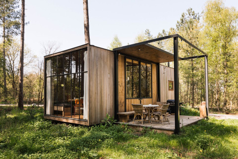Cabaña moderna de madera con grandes ventanales y terraza al aire libre en un claro del bosque primaveral.