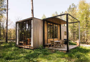 Moderna cabina in legno con ampie finestre e veranda immersa in una radura verdeggiante di bosco primaverile.
