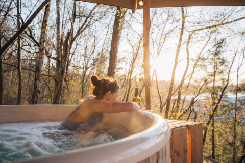 Mujer relajándose en un jacuzzi al aire libre en Cosy Cabins en el bosque de Limburg, Bélgica, al atardecer.