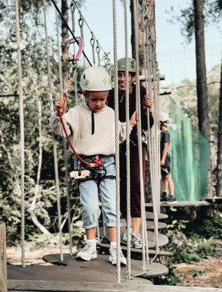 Niño y adulto con cascos cruzan puente colgante en Cosy Cabins, parque de vacaciones en Limburg, Bélgica.