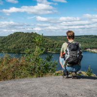 Person mit Rucksack blickt auf einen See und Glamping-Unterkünfte in einem naturnahen Ferienpark.