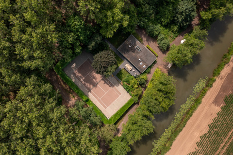 Aerial view of a tennis court and house by a canal, surrounded by forest at Buitengoed Ruysbos, Gelderland.