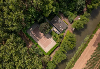 Aerial view of a tennis court and house by a canal, surrounded by forest at Buitengoed Ruysbos, Gelderland.
