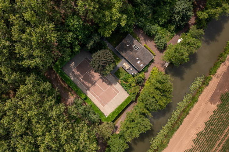Luchtfoto van een tennisbaan en huis aan het water in het bos bij Buitengoed Ruysbos, Gelderland.