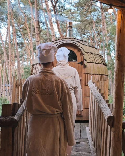 Dos personas con albornoces y toallas caminan hacia una sauna de madera en el bosque de Cosy Cabins Limburg.