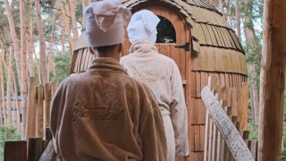 Zwei Personen in Bademänteln und Handtüchern gehen zu einer Holzsauna im Wald bei Cosy Cabins Limburg.