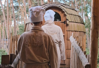 Due persone in accappatoi e asciugamani camminano verso una sauna di legno nella foresta a Cosy Cabins Limburg.