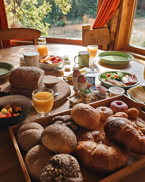 Mesa de desayuno con pan fresco, jugo de naranja, frutas y fiambres junto a una ventana con bosque.