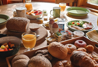 Tavola da colazione con pane fresco, succo d’arancia, frutta e salumi vicino a una finestra sul bosco.