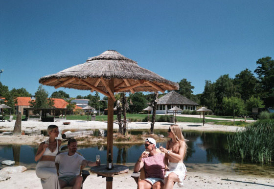 Four people relax under a straw umbrella on the sandy lakeside at Cosy Cabins in Limburg, Belgium.
