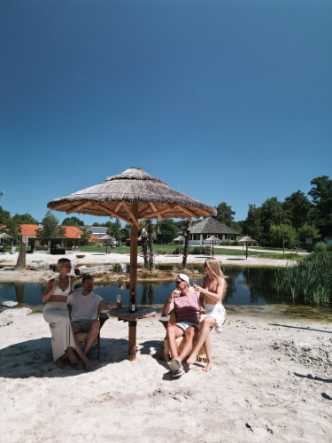 Four people relax under a straw umbrella on the sandy lakeside at Cosy Cabins in Limburg, Belgium.