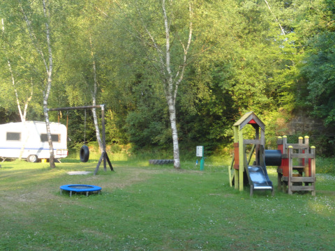 Parque infantil y caravana en Camping Les Bouleaux, Grand Est, Francia, rodeados de árboles verdes.