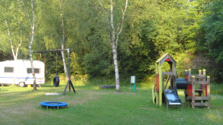 Parque infantil y caravana en Camping Les Bouleaux, Grand Est, Francia, rodeados de árboles verdes.