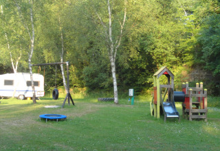 Parque infantil y caravana en Camping Les Bouleaux, Grand Est, Francia, rodeados de árboles verdes.