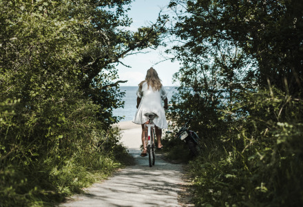 Een vrouw in een witte jurk fietst op een pad omringd door bomen richting het strand bij een vakantiepark.