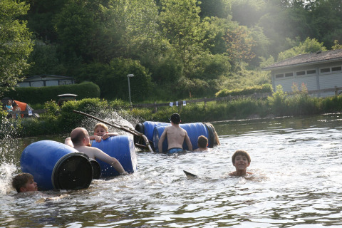 Kinderen en volwassenen zwemmen en spelen met vlotten van blauwe tonnen in een rivier bij Camping Donnersberg.